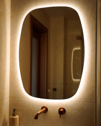 A modern oval bathroom mirror with a warm white LED light border, mounted on a beige tiled wall above a sink with a wall-mounted faucet.